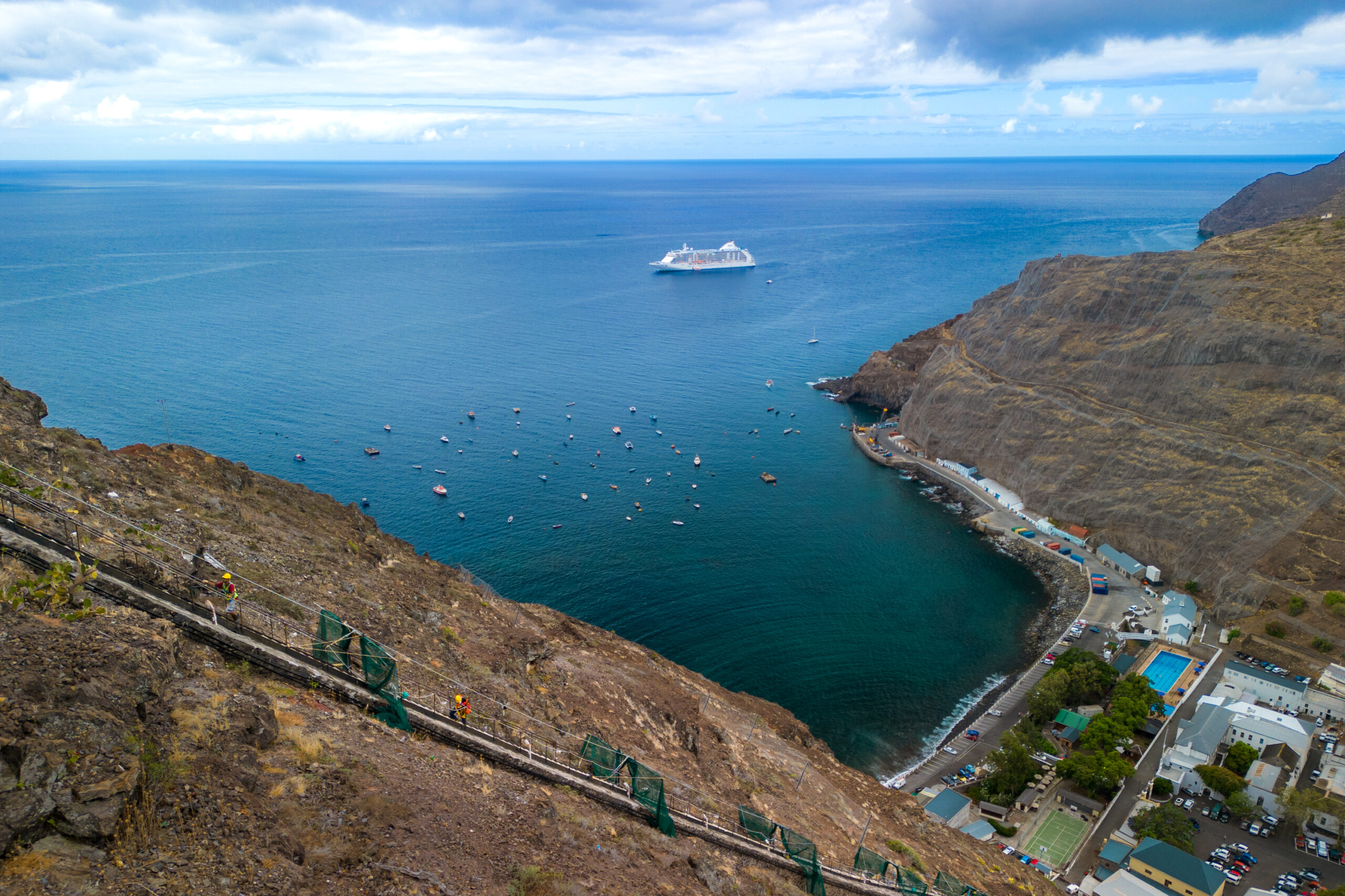 A cruise ship floats near a rocky coastline with small boats and a town by the water under a partly cloudy sky.